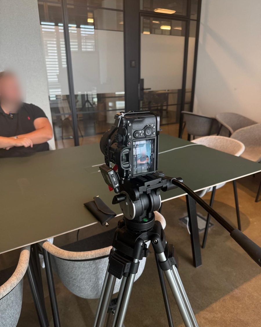 Kamera auf Stativ, im Hintergrund der Interviewpartner am Tisch